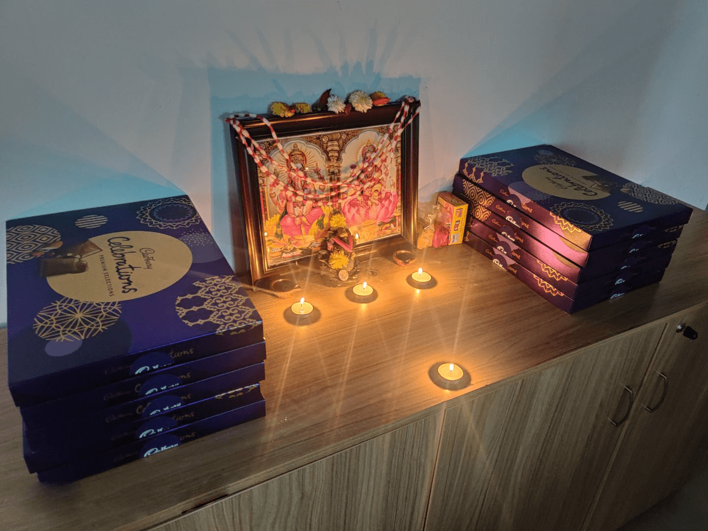 A small prayer setup with candles lit in front of a framed deity picture, surrounded by stacks of celebration gift boxes.